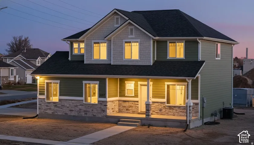 Two-story Colonial-style home featuring a welcoming front porch with stone base and columns, illuminated windows throughout the upper and lower levels, and a clean driveway approach