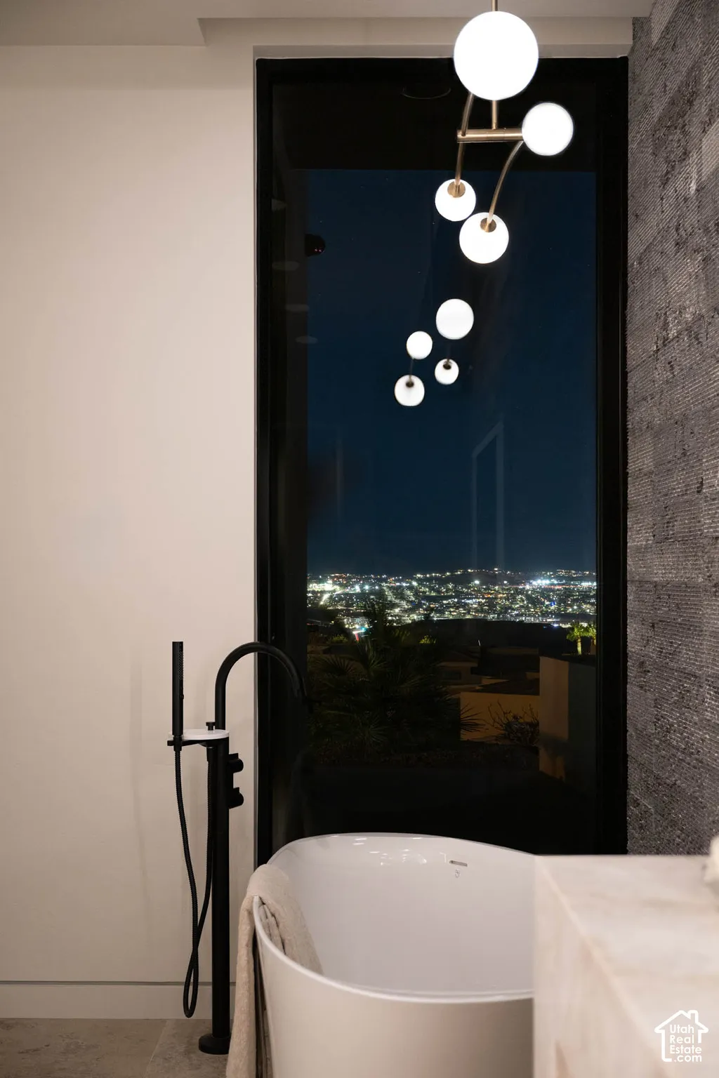 Freestanding soaking tub with a matte black floor-mounted faucet, large floor-to-ceiling window, textured wall accent, and modern spherical pendant lighting