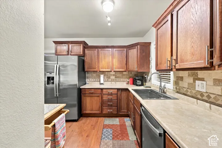 Kitchen featuring natural wood cabinetry, stainless steel appliances, neutral stone-finish countertops, decorative tile backsplash, and wood-finish flooring
