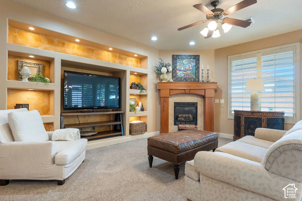 Built-in media center with recessed lighting and shelving, wood-surround fireplace with a hearth, ceiling fan with integrated lighting, window with plantation shutters, and light-toned carpet flooring