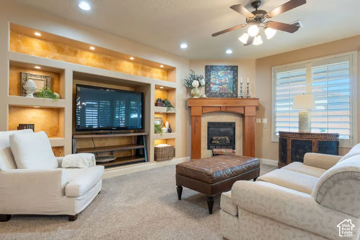 Built-in media center with recessed lighting and shelving, wood-surround fireplace with a hearth, ceiling fan with integrated lighting, window with plantation shutters, and light-toned carpet flooring