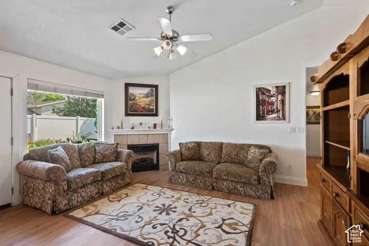 Vaulted ceiling with a ceiling fan, fireplace with a tile surround and mantel, large window, and wood-finish flooring