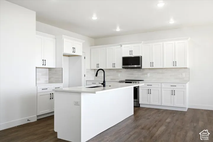 White shaker-style cabinetry with black hardware, a center island with a sink and matte black faucet, light-toned countertop, and wood-finish flooring