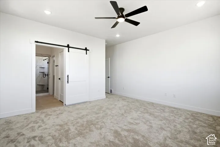 Spacious room featuring neutral carpeting, a ceiling fan with integrated lighting, recessed lighting, and a modern barn door entry to an adjacent bathroom with a glass-enclosed shower