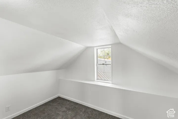 Sloped ceiling room featuring a single-hung window, recessed wall, and dark gray carpeting