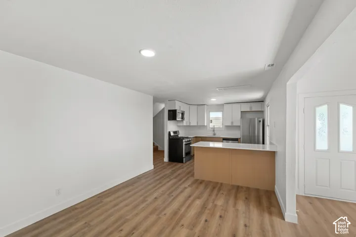 Integrated kitchen and living area featuring wood-finish flooring, recessed lighting, white cabinetry, a kitchen island with a light-toned countertop, and stainless steel appliances