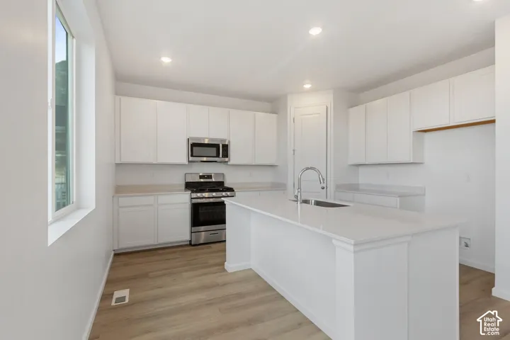 White cabinetry, stainless steel appliances, kitchen island with sink and faucet, recessed lighting, and wood-finish flooring