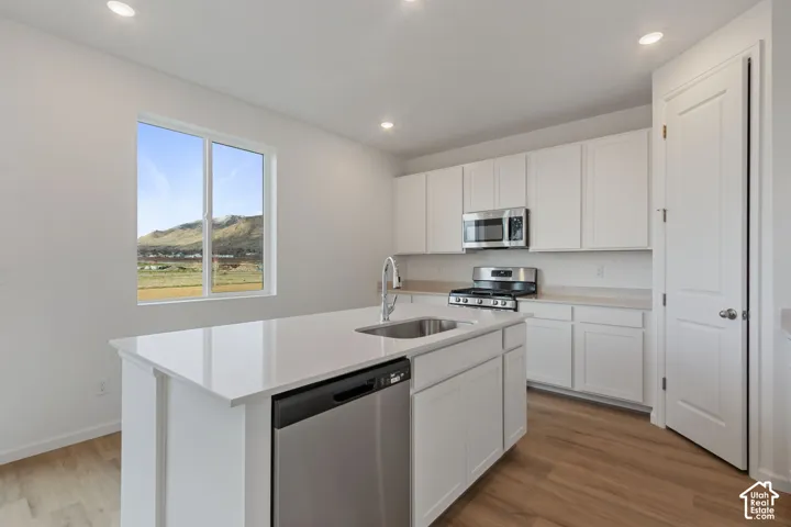 Kitchen featuring white cabinetry, stainless steel appliances, a center island with sink, quartz countertops, and wood-finish flooring