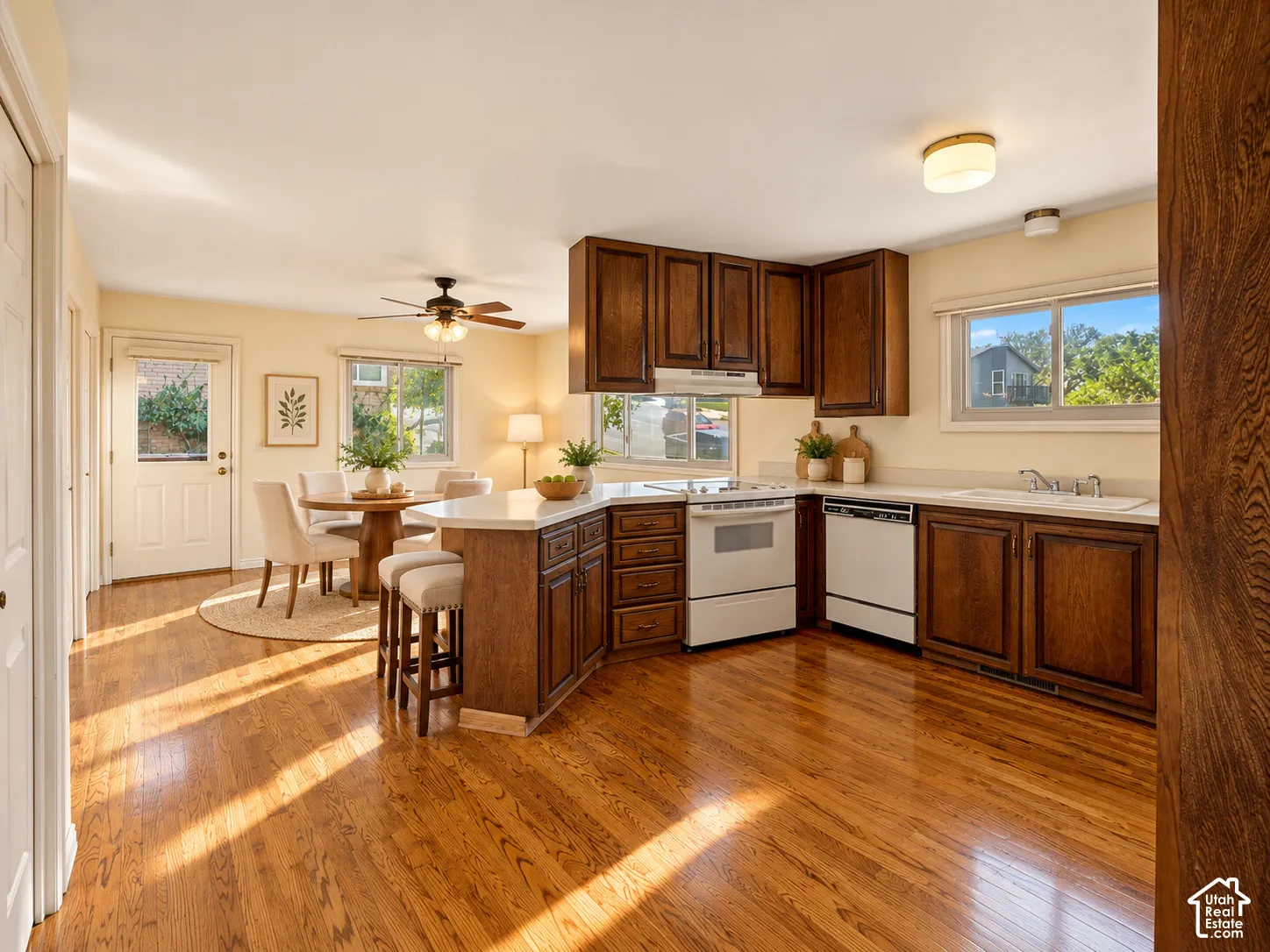 Hardwood flooring throughout, L-shaped kitchen island with seating, dark wood cabinetry, white appliances, and a ceiling fan