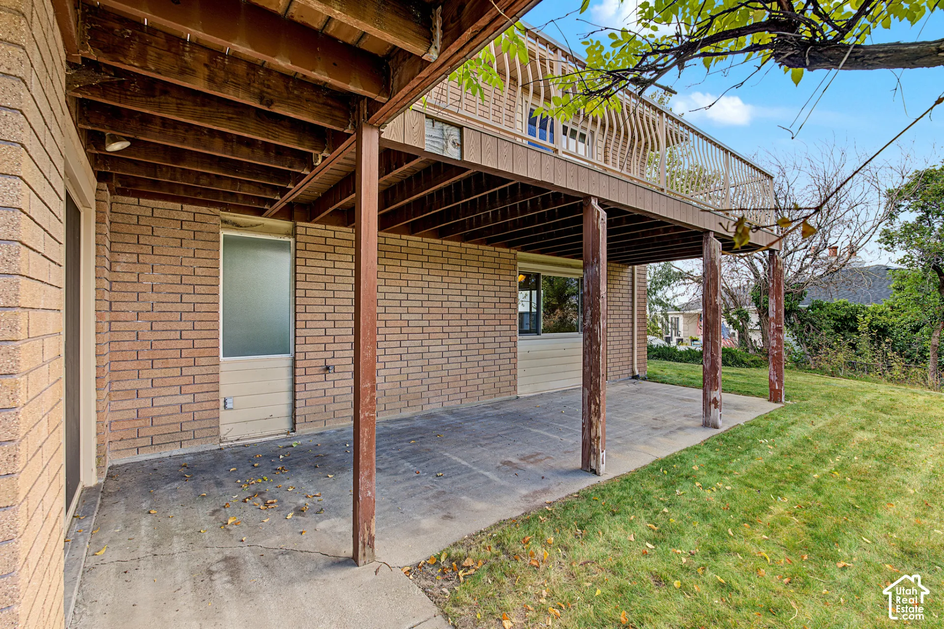 Expansive concrete patio with overhead deck structure, supported by wood posts, featuring brick exterior walls and a grassy area