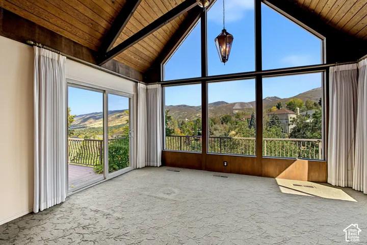 Vaulted wood-paneled ceiling, exposed wood beams, expansive floor-to-ceiling windows, sliding glass door access to an exterior deck, and patterned carpeting