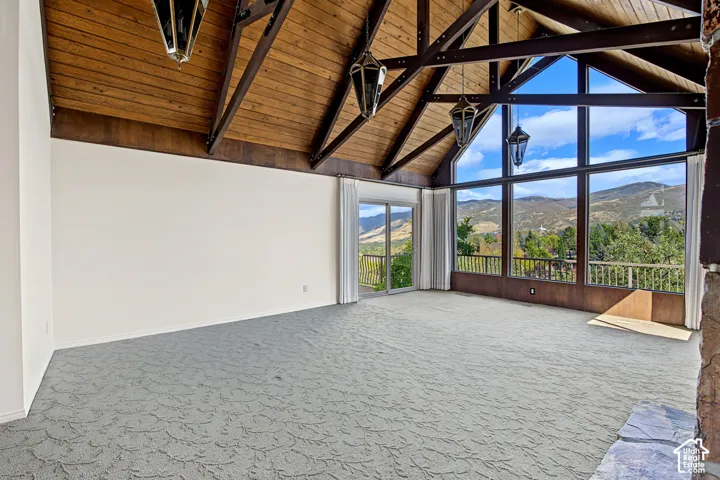 Vaulted wood-paneled ceiling with exposed beams, expansive floor-to-ceiling windows, sliding glass door leading to a balcony, and a stone hearth