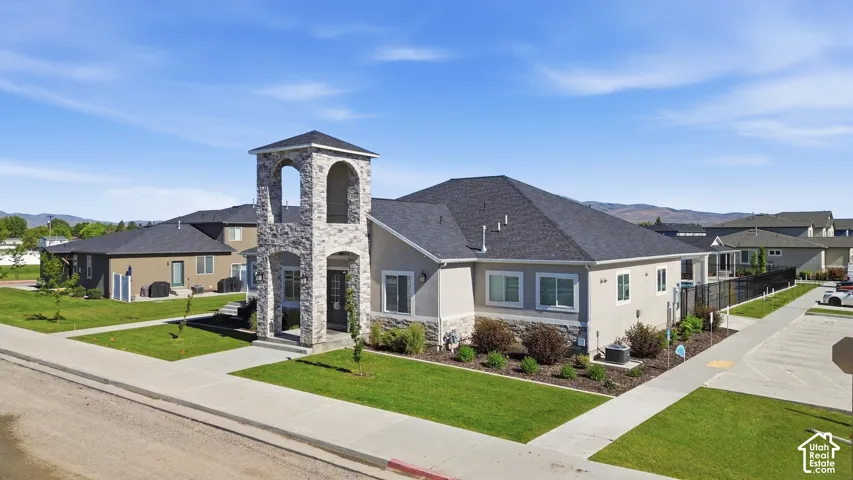 Stone-clad entryway tower, stucco exterior, dark shingle roofing, landscaped grounds, and paved parking area