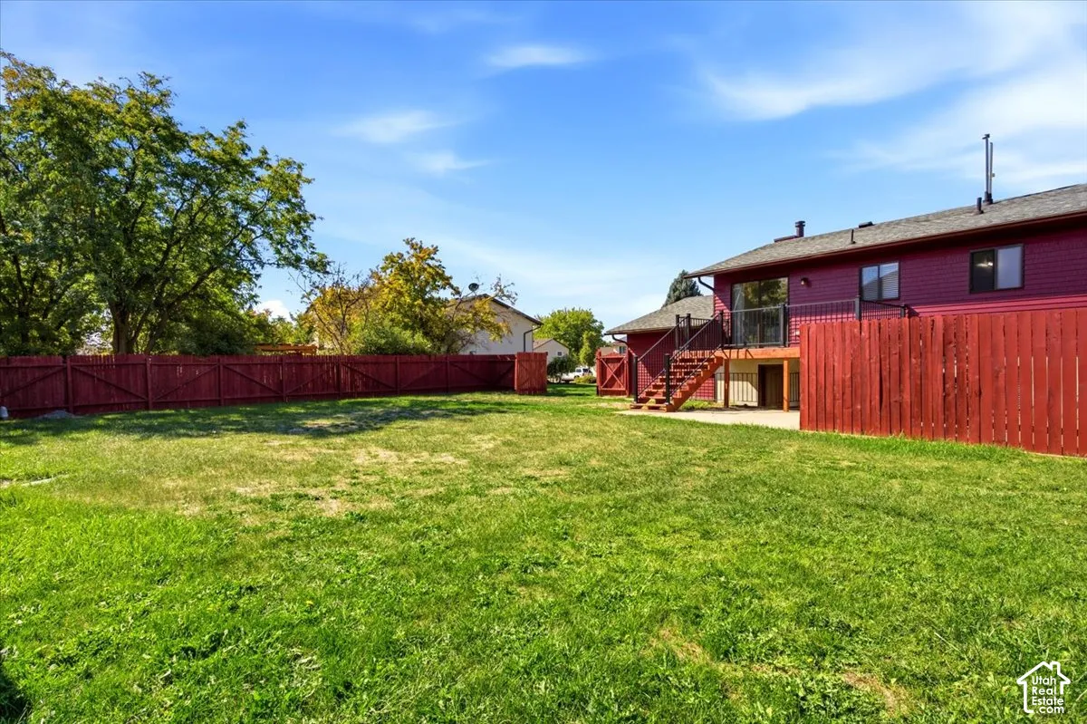Expansive grassy yard with mature perimeter trees, featuring a red wood privacy fence and a two-story exterior with a deck and lower-level patio