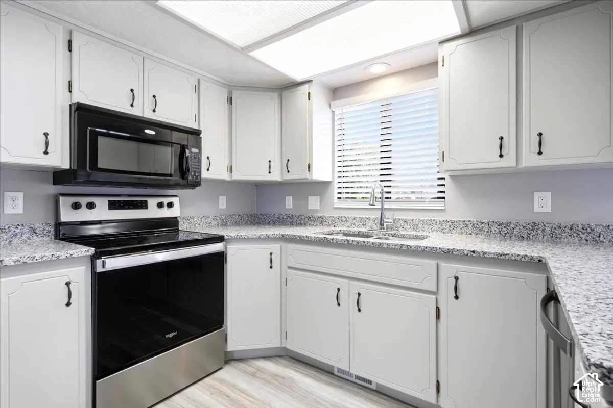 White cabinetry with dark hardware, stone-finish countertops, stainless steel appliances, stainless steel sink with a high-arch faucet, and wood-finish flooring
