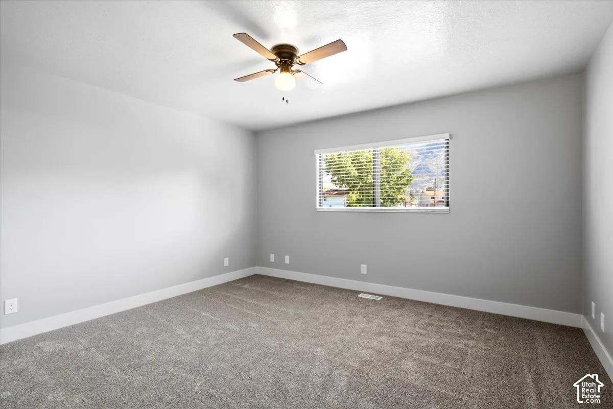 Neutral gray walls, textured carpet flooring, ceiling fan with light fixture, and a window with blinds