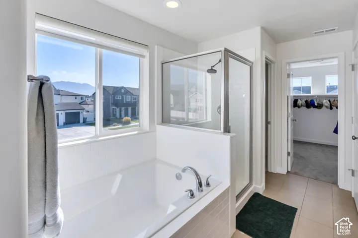 Recessed lighting, large window with roller shades, built-in bathtub with deck-mounted faucet, glass-enclosed shower with subway tile surround, and tile flooring