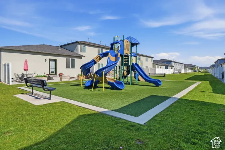 Outdoor recreational area featuring a playground structure with slides and climbing elements, surrounded by artificial turf and natural grass, with concrete pathways and benches