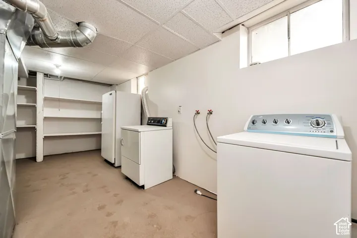 Spacious laundry room featuring white front-load washer and dryer units, built-in shelving along the perimeter, concrete flooring, and natural light from high windows
