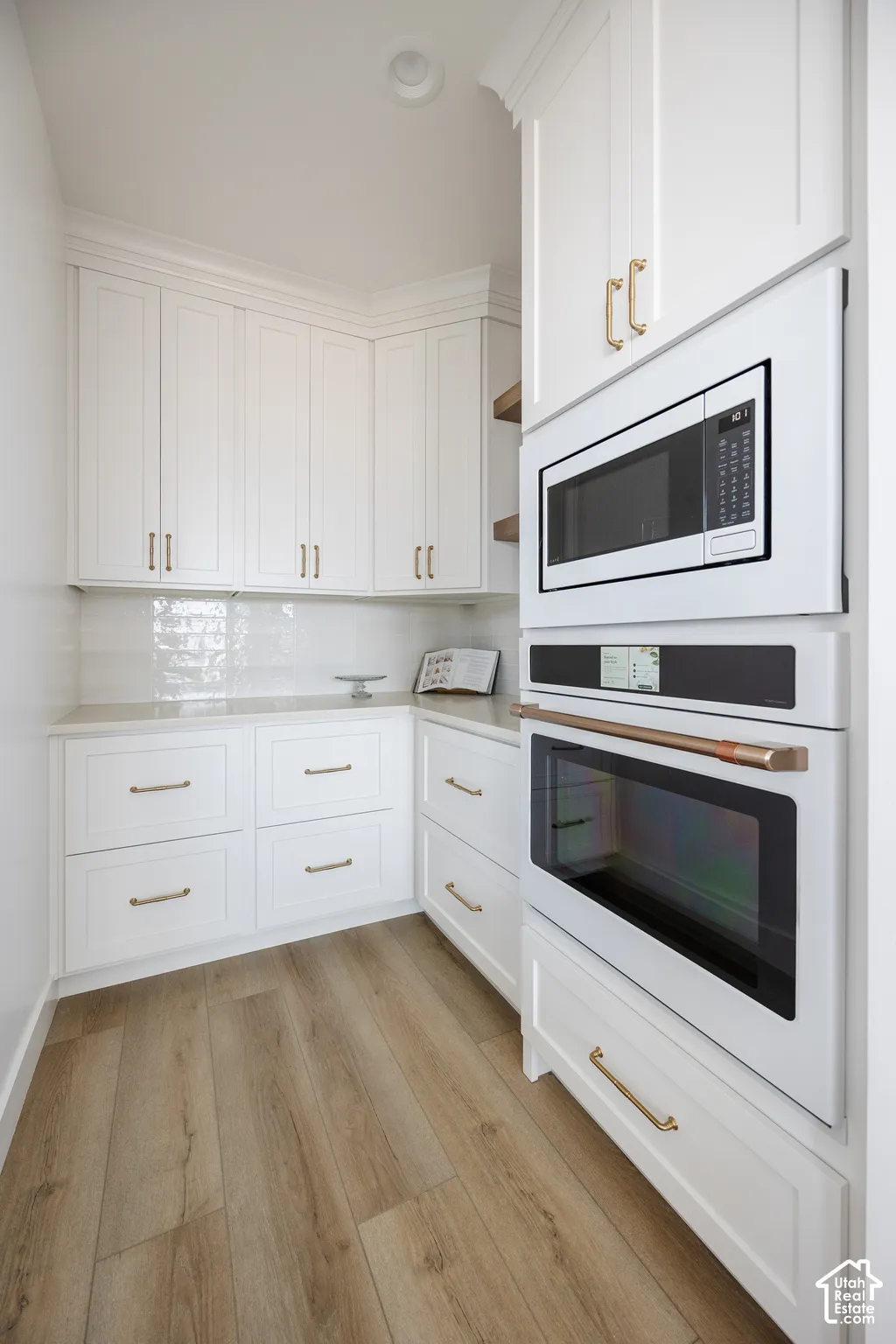 White cabinetry with gold-tone hardware, wood-finish flooring, built-in microwave, built-in oven, and light-toned countertops