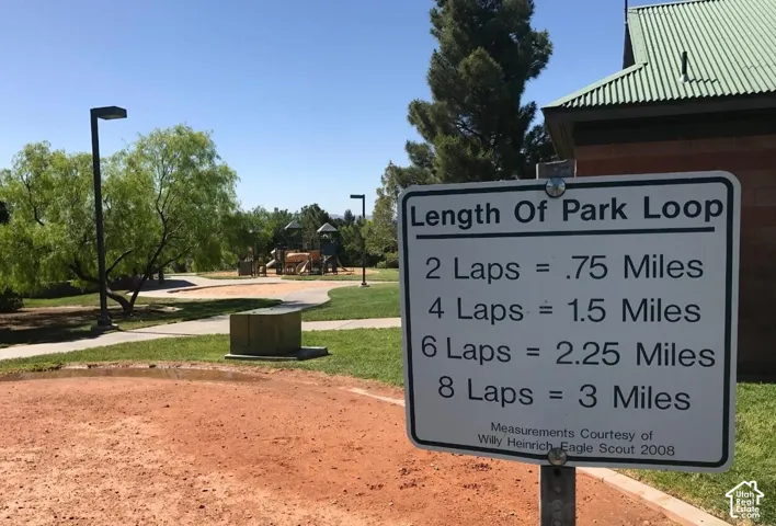 Paved walking path, green grass landscaping, mature trees, playground structures, and a park building with a metal roof