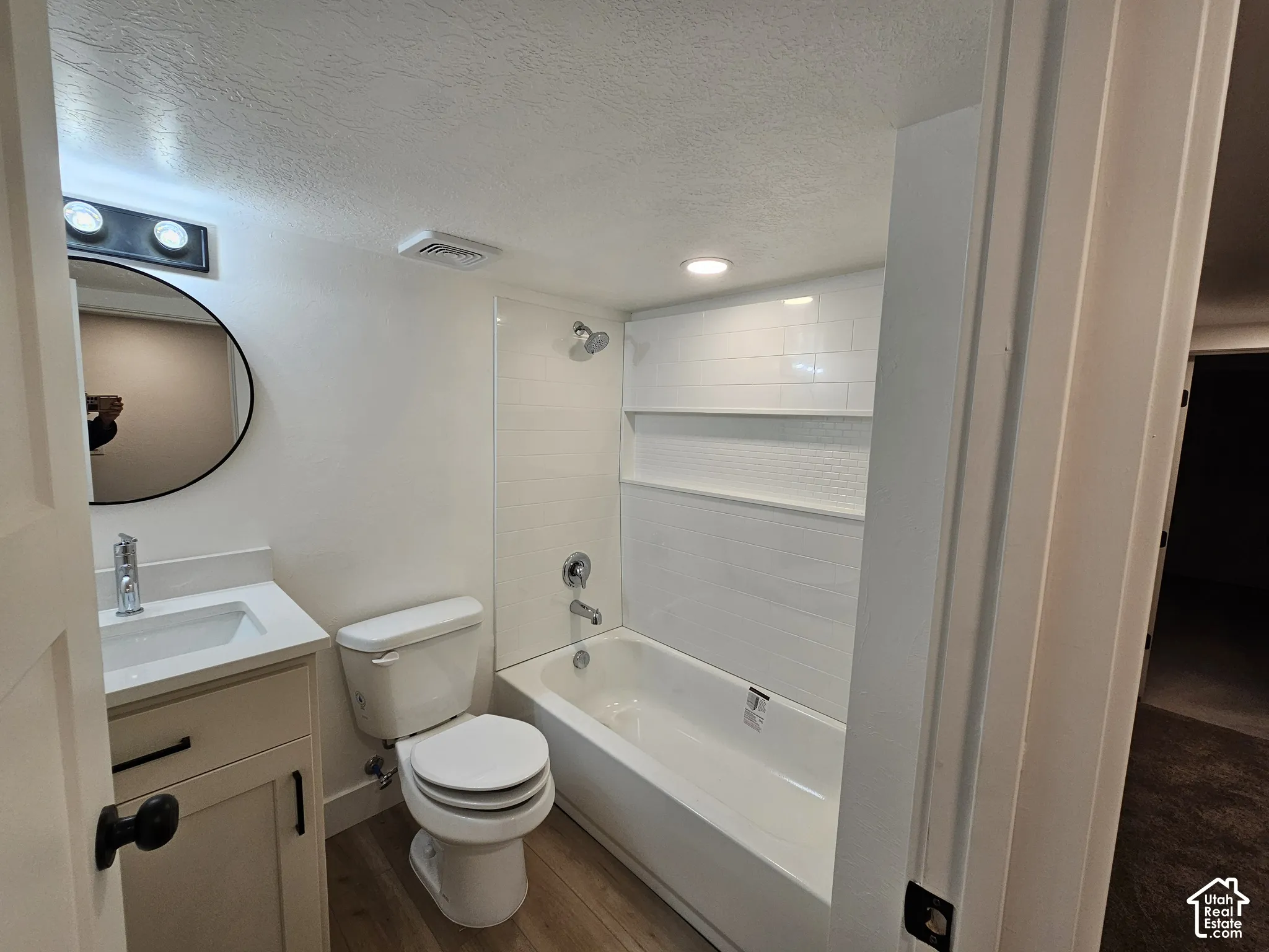Bathroom featuring a white vanity with an integrated sink, a circular mirror, a built-in tub with white subway tile surround, and wood-finish flooring