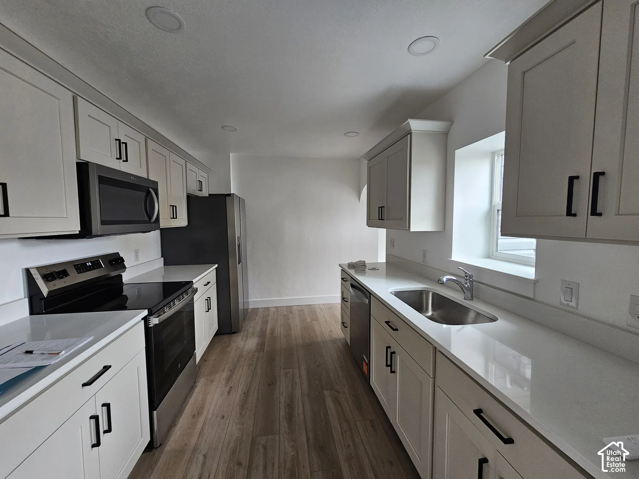 White shaker-style cabinetry with matte black hardware, stainless steel appliances, light-toned countertops, wood-finish flooring, and recessed lighting