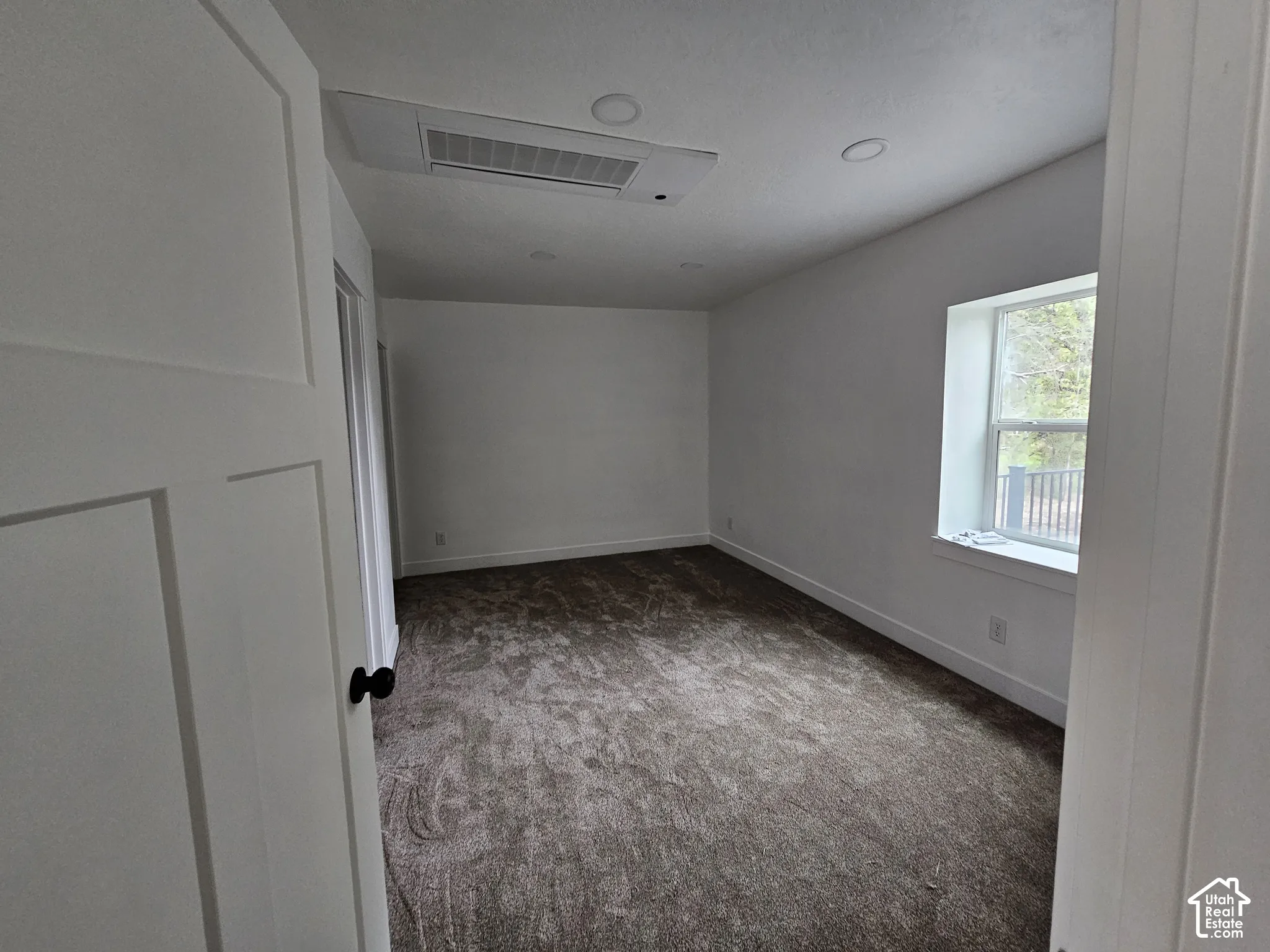 White interior walls, recessed ceiling lighting, large window with white trim, dark textured carpeting, and white interior door with black hardware