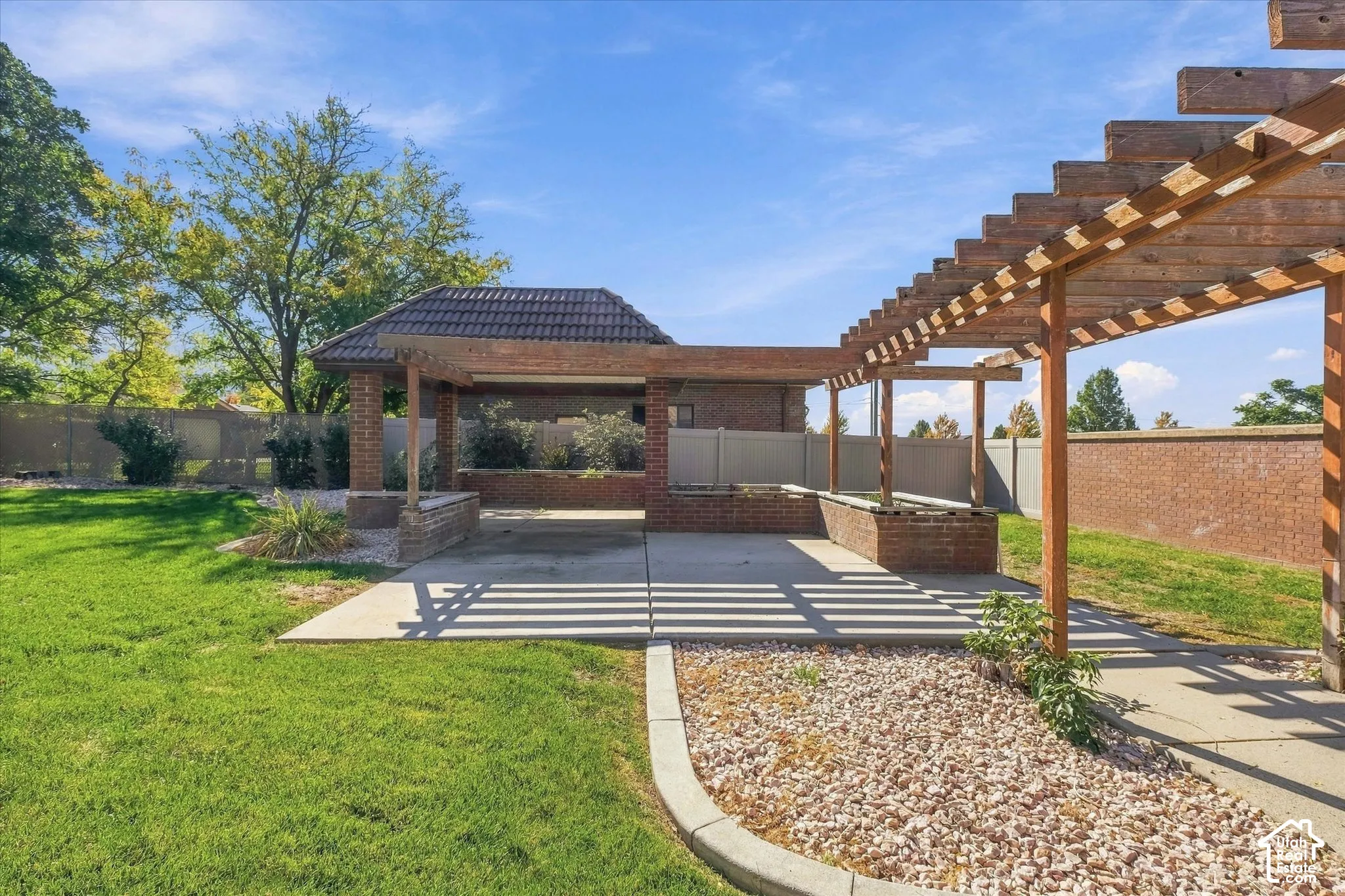 Expansive outdoor space featuring a covered patio with a brick-built structure, a long wooden pergola, concrete hardscaping, and a manicured lawn