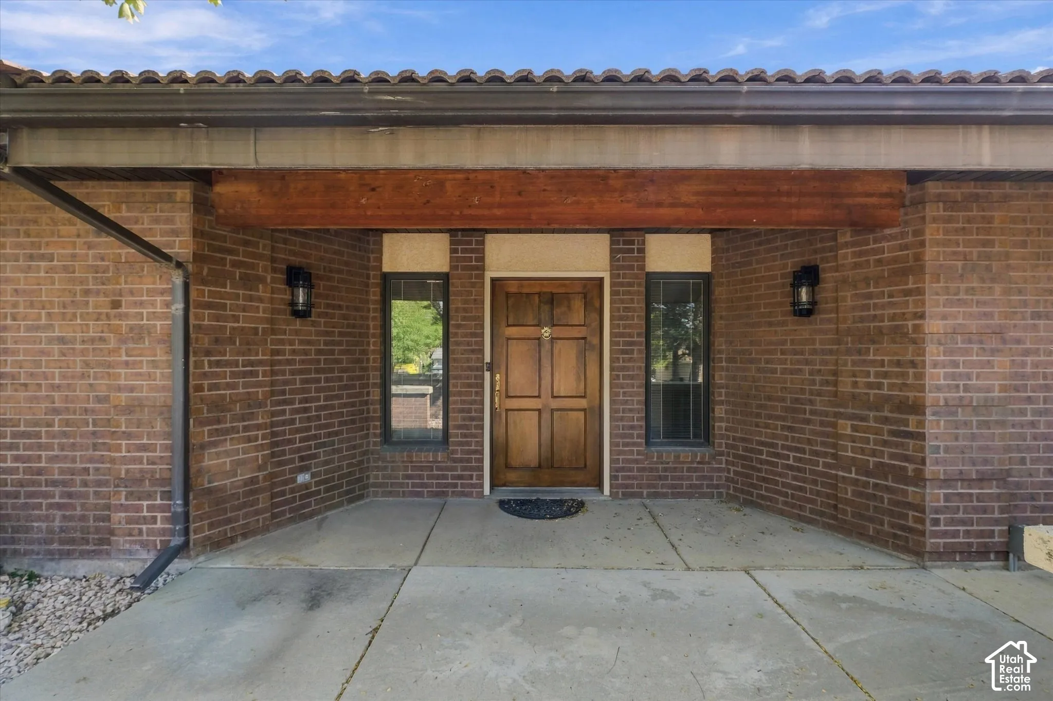 Brick exterior featuring a wooden entry door, sidelight windows, overhead wood-finish beam, and exterior sconces