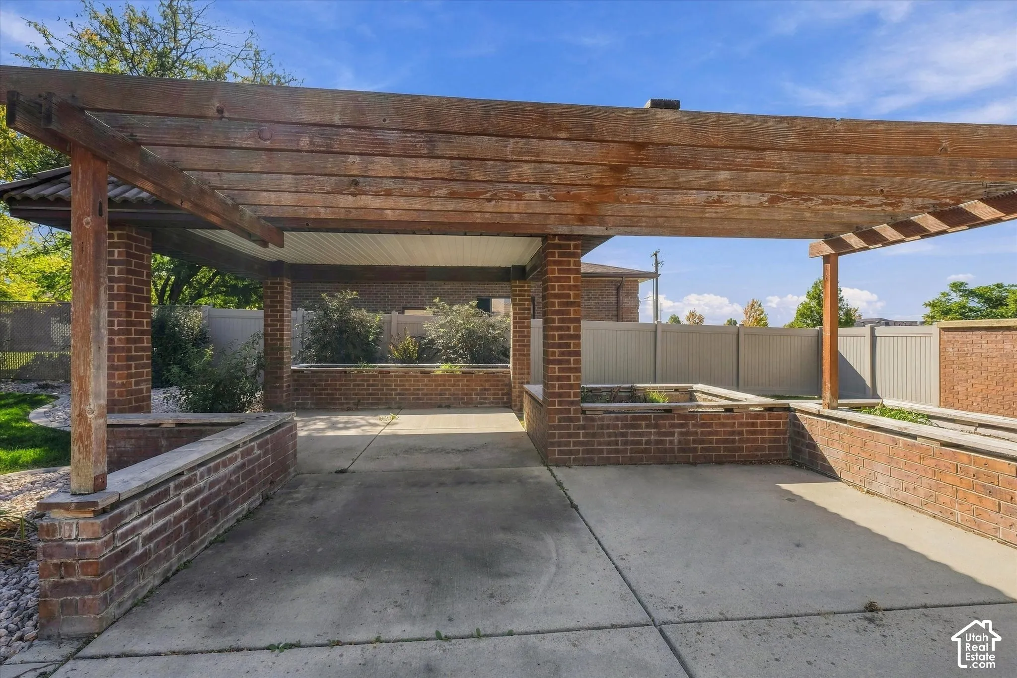 Expansive outdoor area featuring concrete paving, a wood-beam pergola, brick-clad planters, and a privacy fence