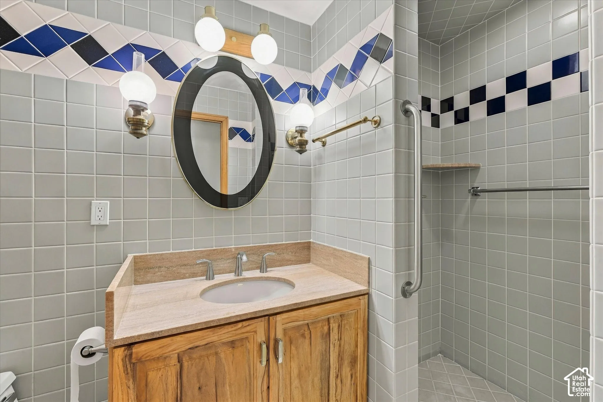 Elegant bathroom featuring soft gray subway tile walls with navy blue accent detailing, wood-finish vanity with dual-sink configuration and light wood countertop, round black-framed mirror, brass fixtures and hardware, and a walk-in shower with glass encl