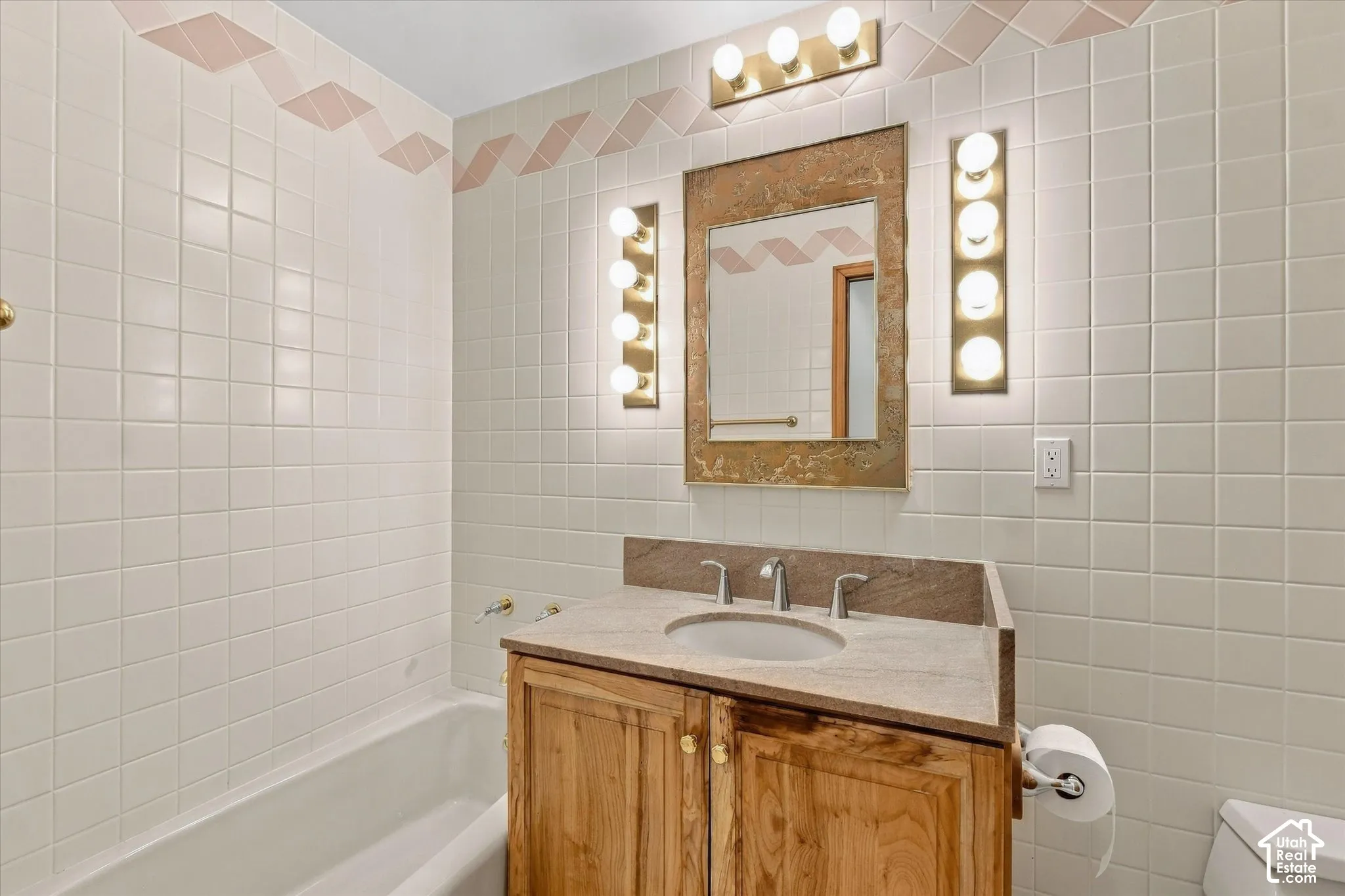 White tiled walls with a decorative border, a vanity with a light-colored countertop and integrated sink, a wood-finish cabinet, and a framed mirror with vanity lighting
