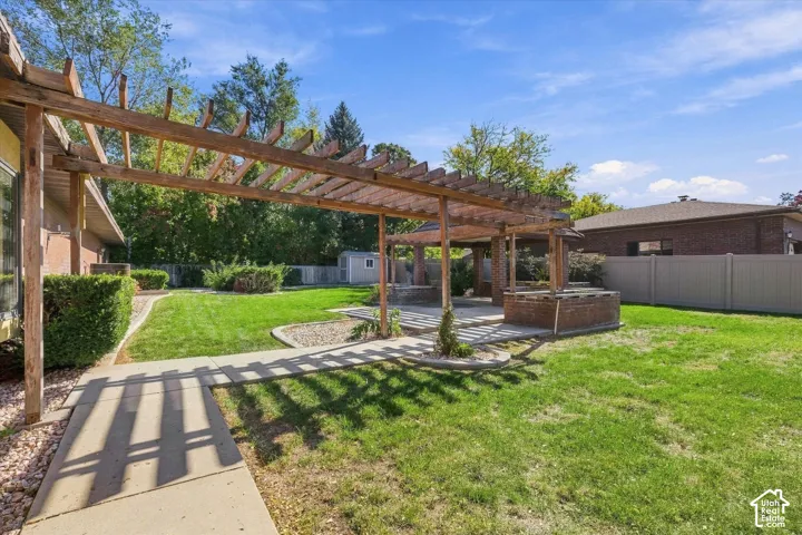 Expansive backyard featuring a large pergola, concrete patio, built-in brick planters, a manicured lawn, and mature shrubs