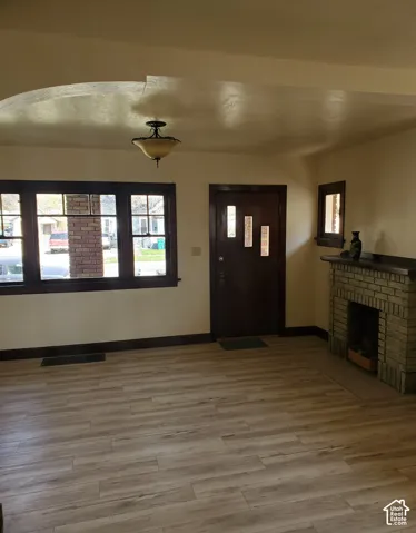 Wood-finish flooring throughout, featuring a brick fireplace with a mantel, a dark wood entry door with vertical glass inserts, and a multi-pane window with dark wood trim