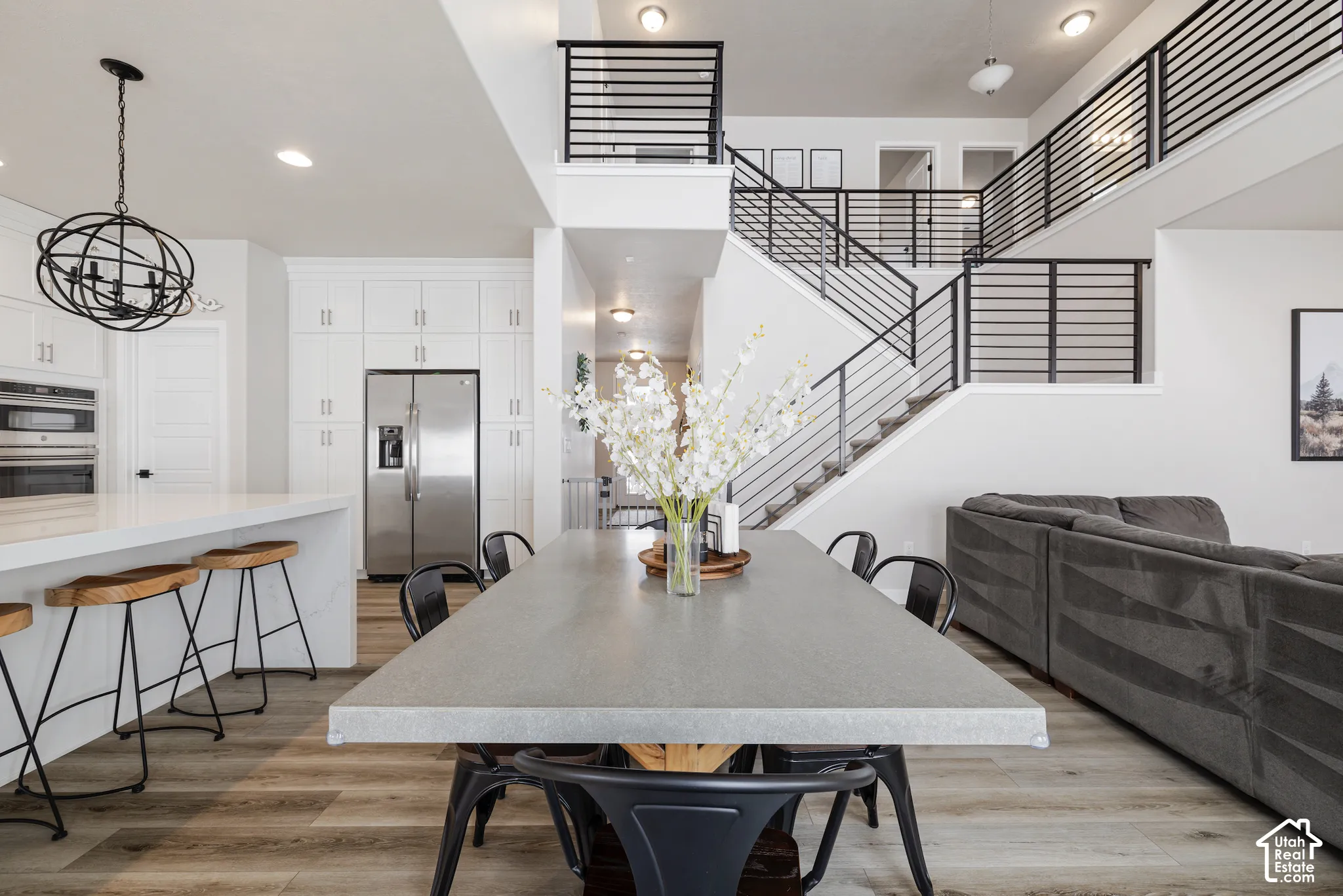 White kitchen cabinetry with stainless steel appliances, wood-finish flooring, a two-story staircase with horizontal metal railings, and a large dining table