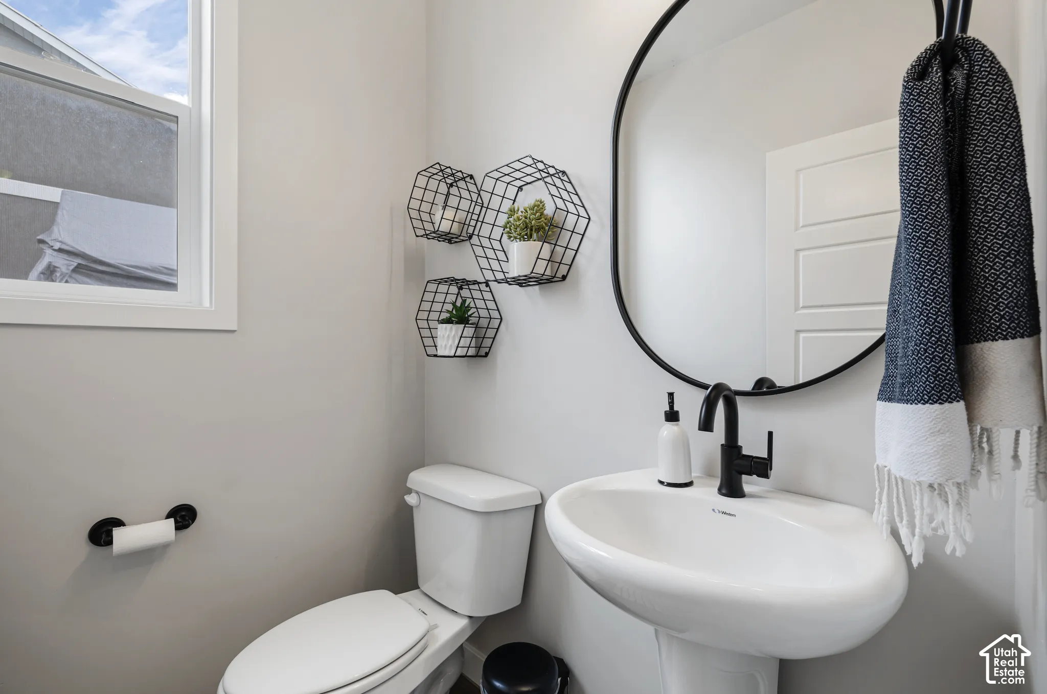 Pedestal sink with a matte black faucet, wall-mounted oval mirror, and a window providing natural light