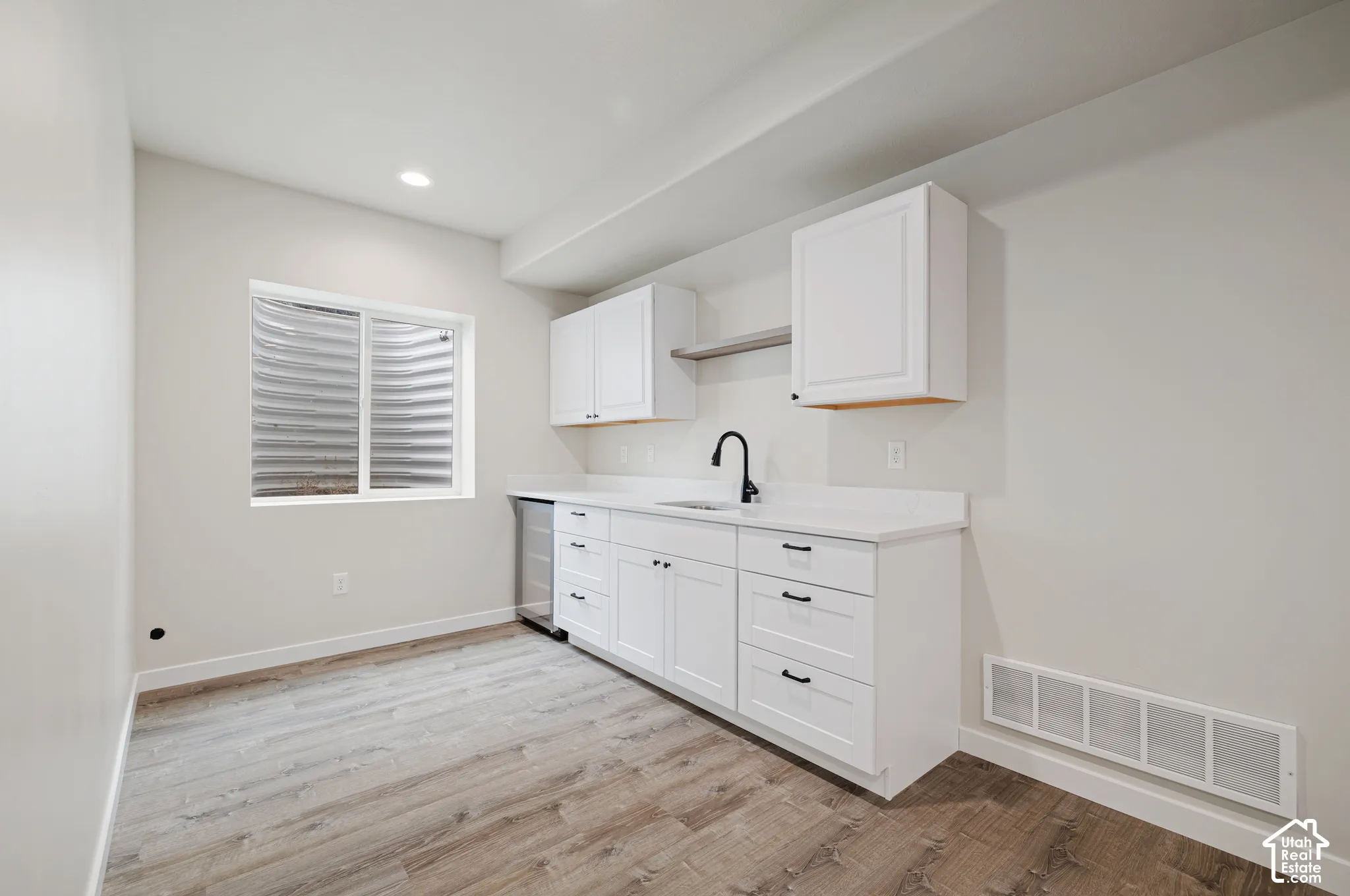 White shaker cabinetry with dark hardware, a stainless steel beverage refrigerator, a single basin sink with a matte black faucet, light-toned countertops, and wood-finish flooring
