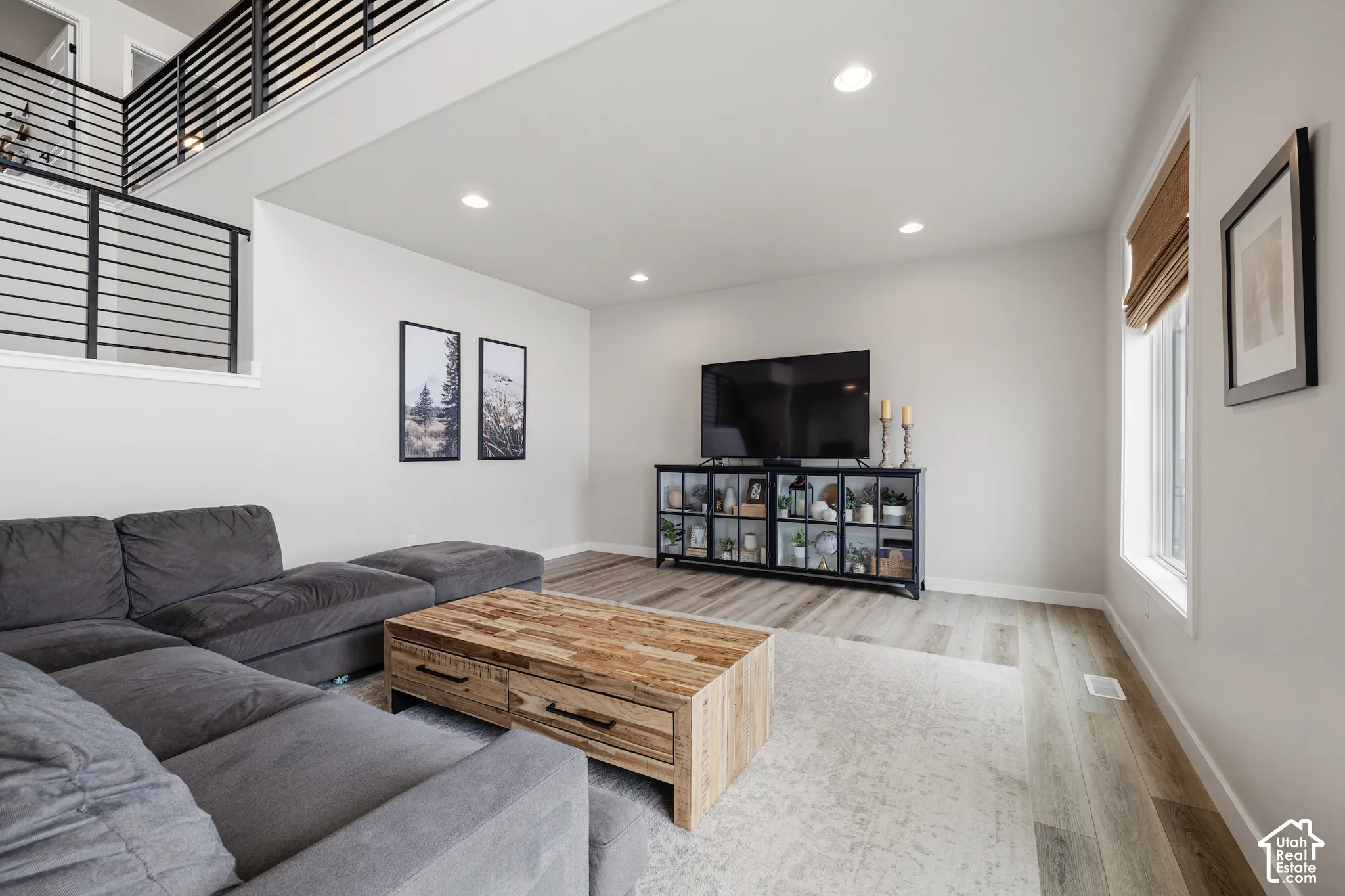 Spacious living area featuring wood-finish flooring, recessed lighting, a modern horizontal metal railing, and a window with a roller shade