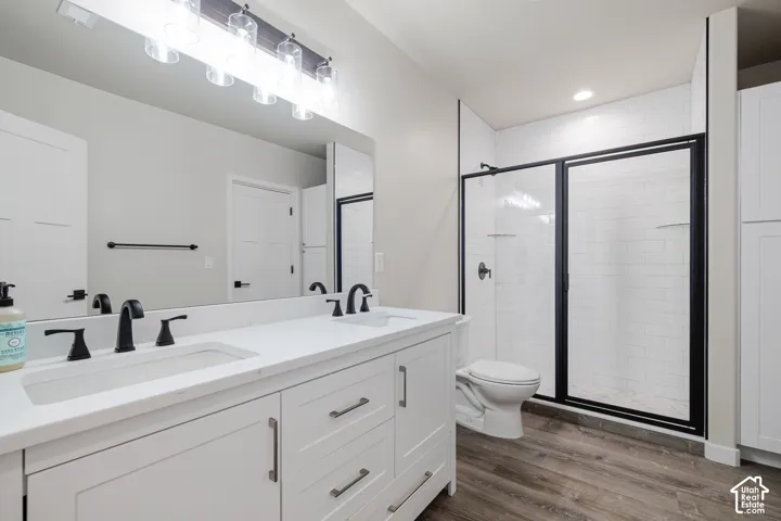 Dual vanity with white cabinetry and matte black faucets, large frameless mirror, glass enclosed shower with subway tile, wood-finish flooring, and recessed lighting