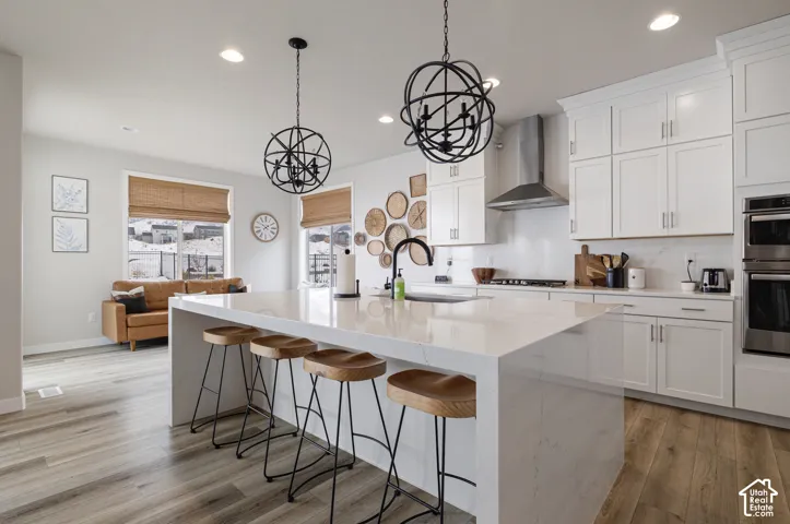 Spacious kitchen featuring white cabinetry, a large waterfall island with seating, a stainless steel range hood, and integrated double ovens