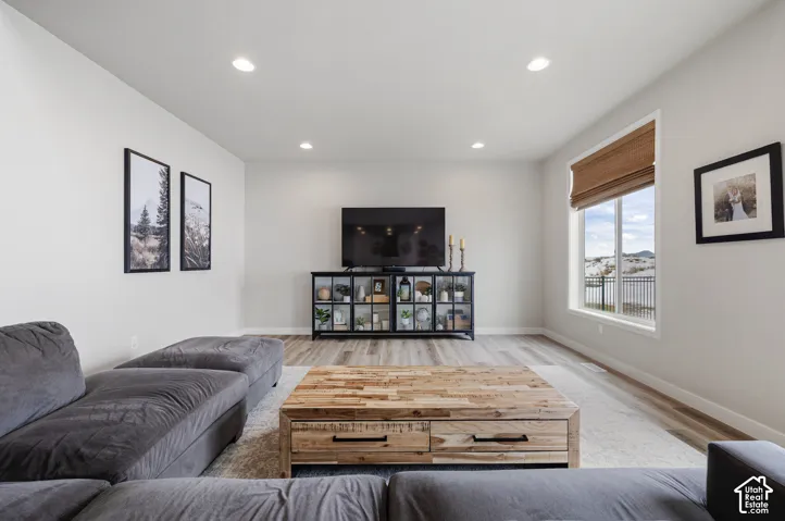 Recessed lighting illuminates the open living space, featuring wood-finish flooring and a large window with a woven shade