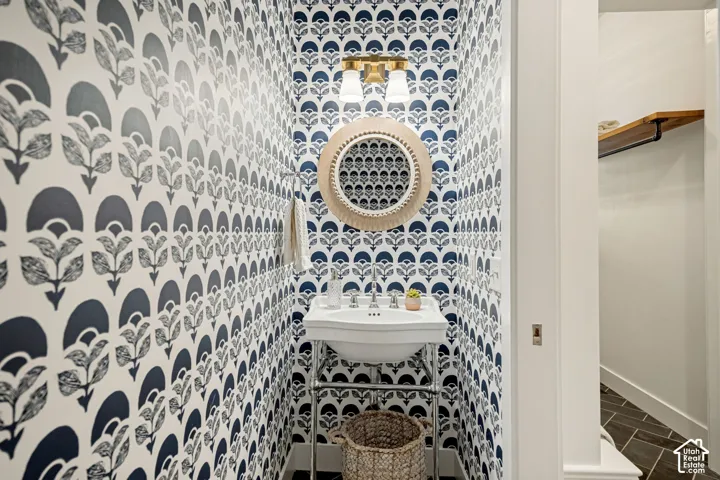 Patterned wallpaper, white pedestal sink with polished chrome stand, circular mirror, brass-finish sconce, and dark tile flooring