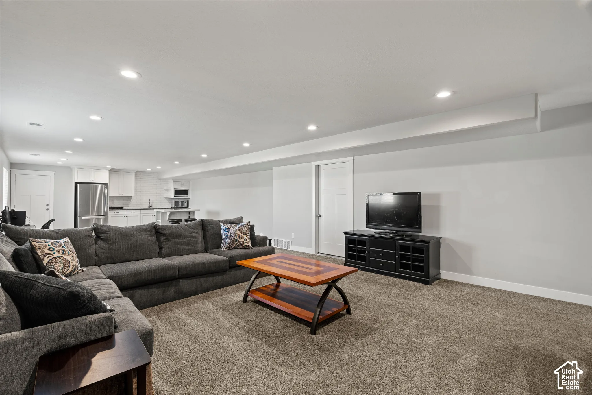 Recessed lighting, light gray walls, white trim, carpeted flooring, and a kitchen area with white cabinetry and a stainless steel refrigerator