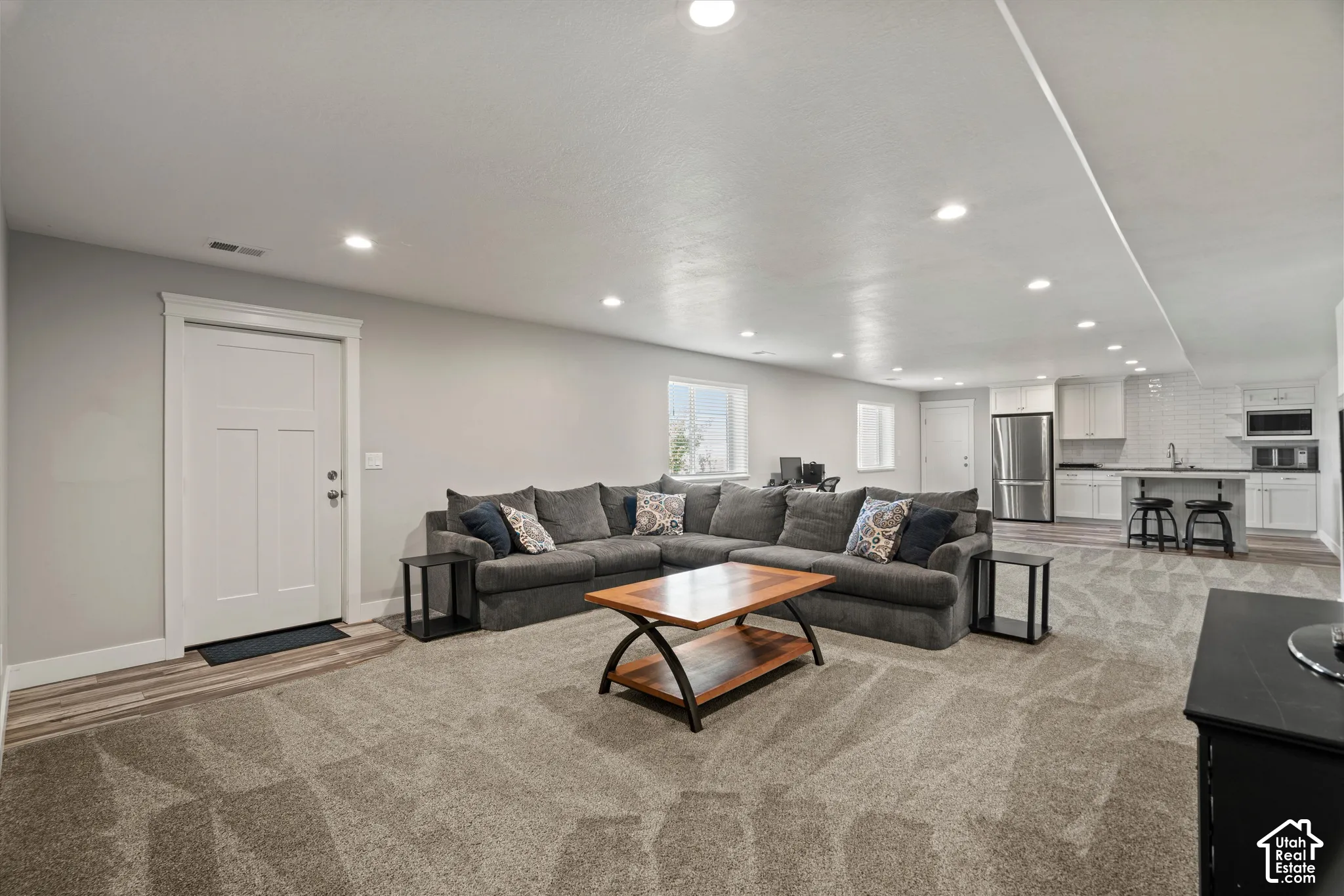 Recessed lighting illuminates the open-concept living space, featuring neutral carpeting and a white trim door