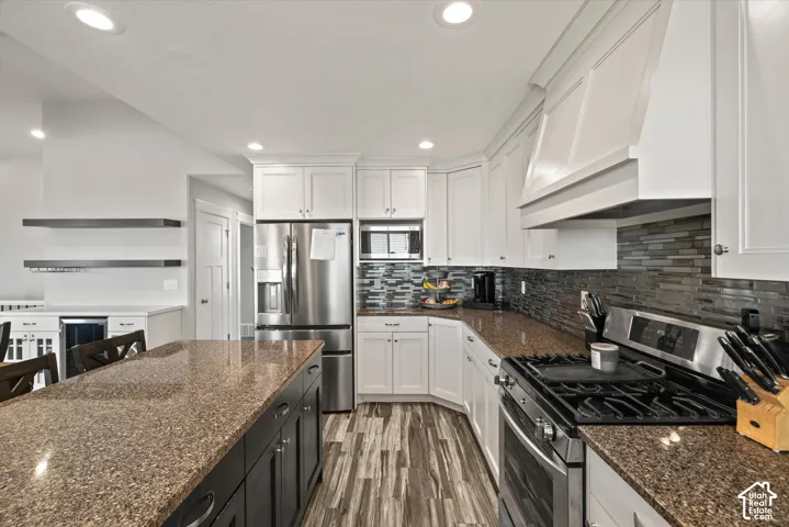 Kitchen featuring white cabinetry, stainless steel appliances, a multi-tone tile backsplash, built-in range hood, and wood-finish flooring