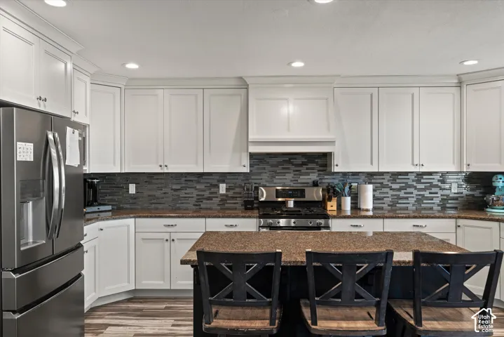 Kitchen featuring white cabinetry, a large kitchen island with counter seating, stainless steel appliances, a mosaic tile backsplash, and wood-finish flooring
