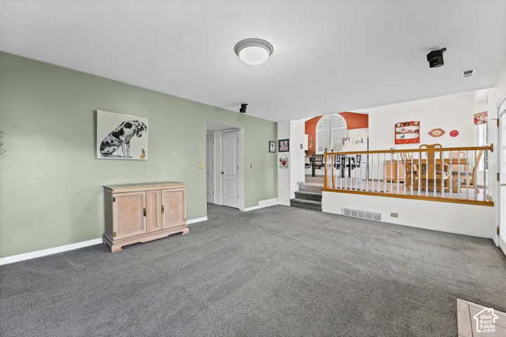 Carpeted living space with a light green accent wall, recessed lighting, and a partial wall with a wood-finished handrail, separating a lower level from a dining area featuring an arched architectural detail