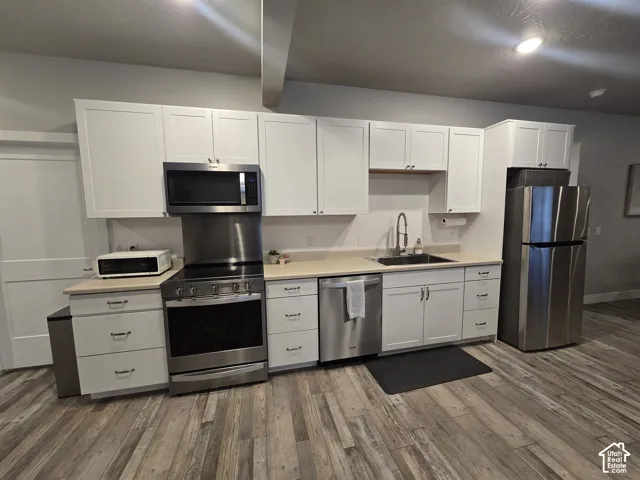 Kitchen featuring white shaker-style cabinetry, stainless steel appliances, a black sink with a gooseneck faucet, light-toned countertops, and wood-finish flooring