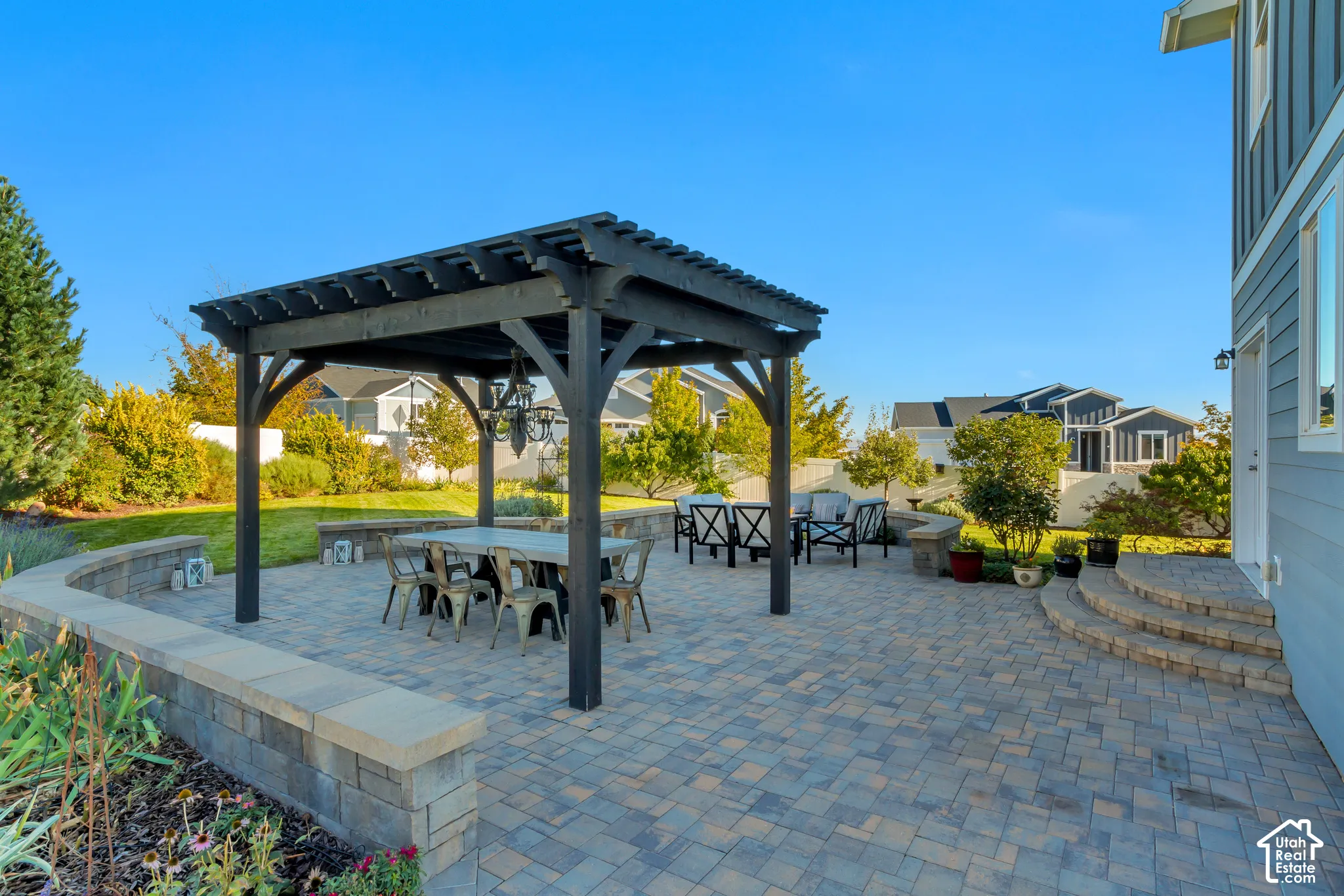 Backyard patio featuring pavers, a Western Timberframe wooden pergola, and a curved stone retaining wall surrounded by beautifully planted garden beds and an expansive grass lawn
